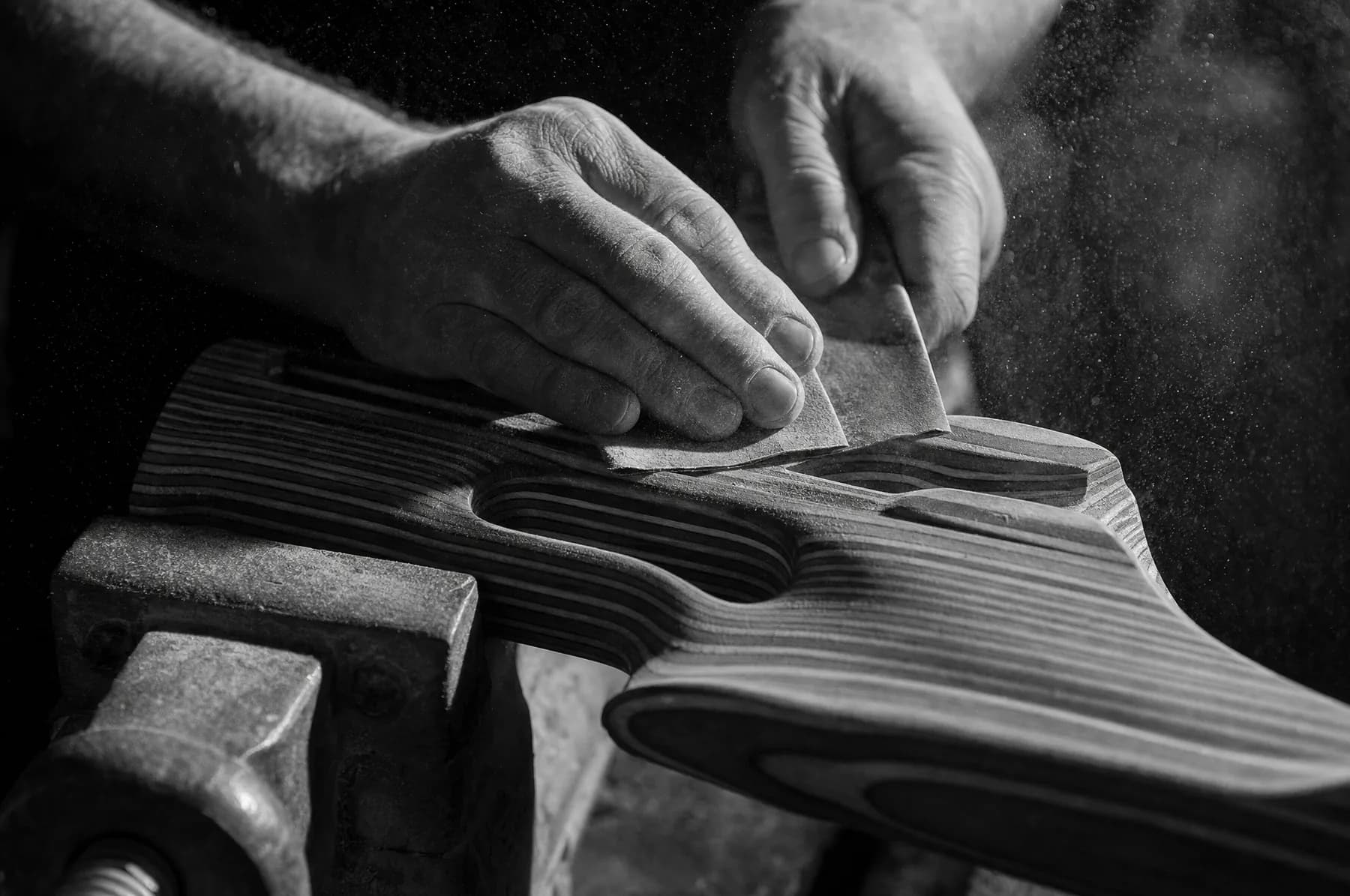 Hands working abrasive paper along the comb of a laminated hardwood stock held in a bench vice, dust hanging in the light