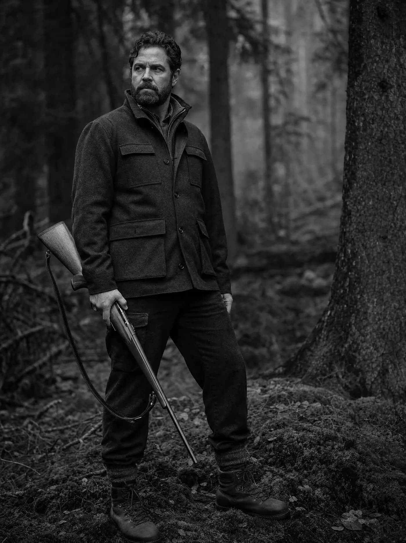 A man in a wool jacket standing in conifer forest, a wood-stocked rifle at his side